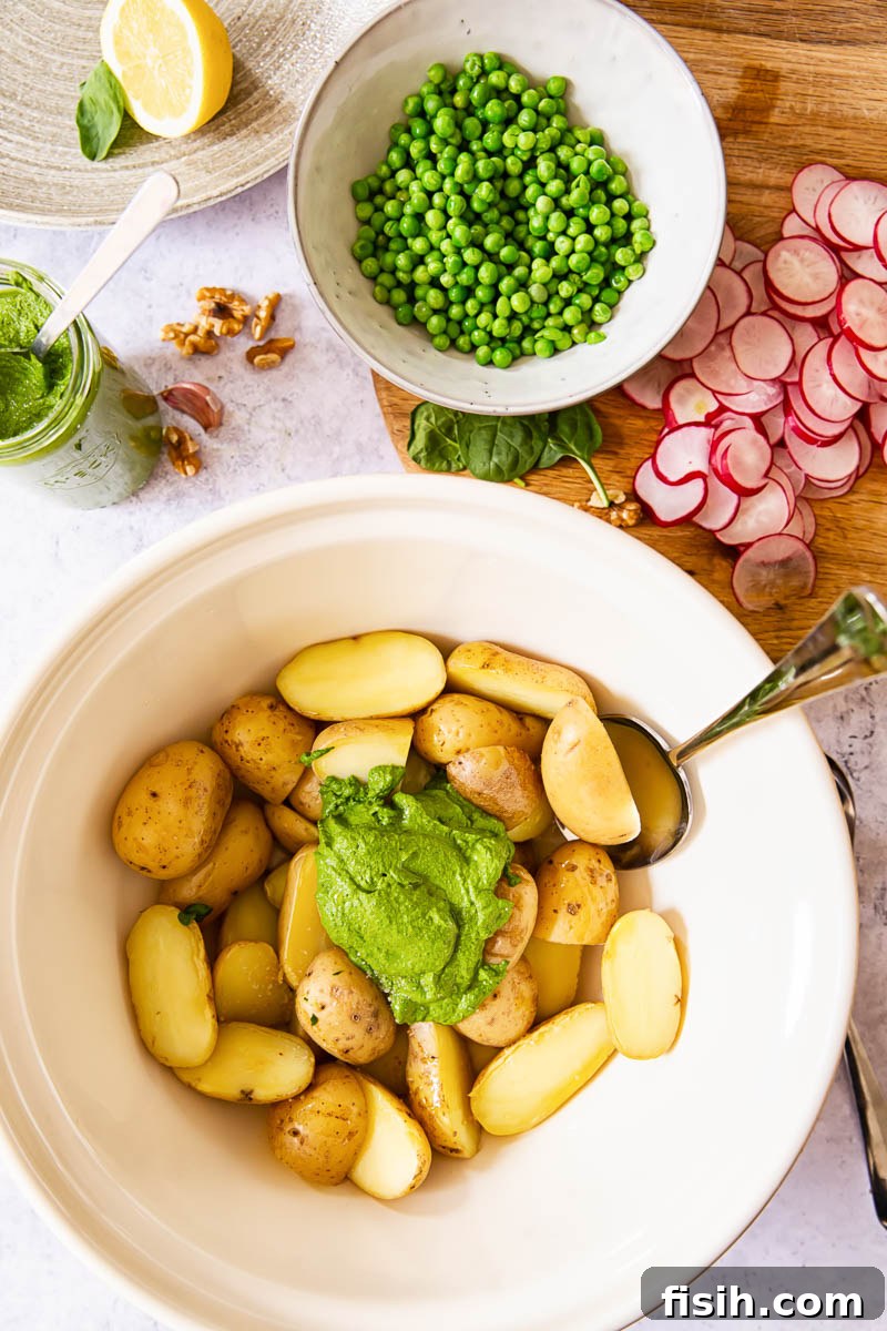 A flat lay of fresh ingredients for Pesto Potato Salad, including small baby potatoes, vibrant radishes, green peas, and a bowl of homemade spinach pesto.