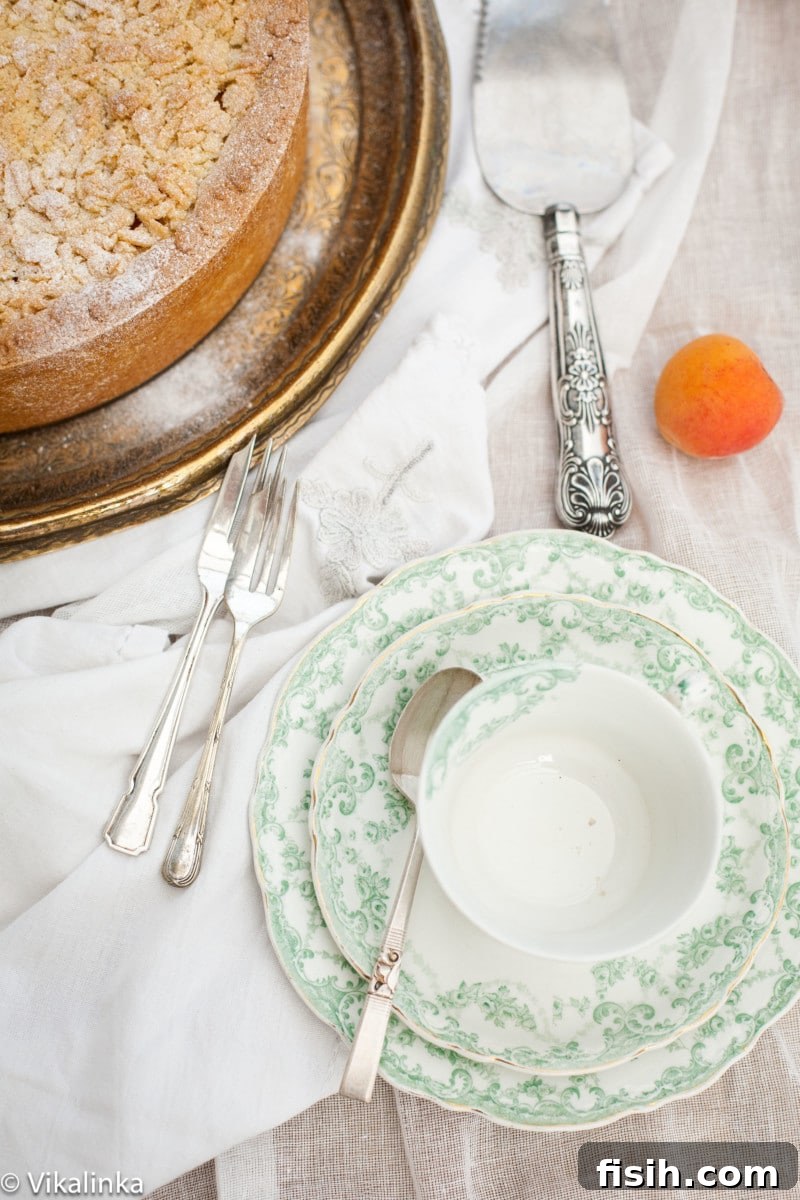 Top-down shot of a teacup on a white tablecloth with a slice of apricot crumb cheesecake in the corner, suggesting a cozy dessert moment.