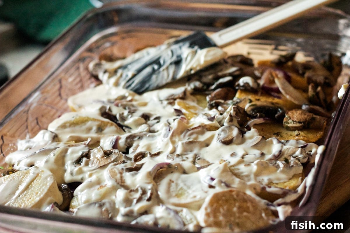 A close-up, top-down view of the layered potatoes, mushrooms, and creamy sauce within a baking dish, before the final cheese topping.