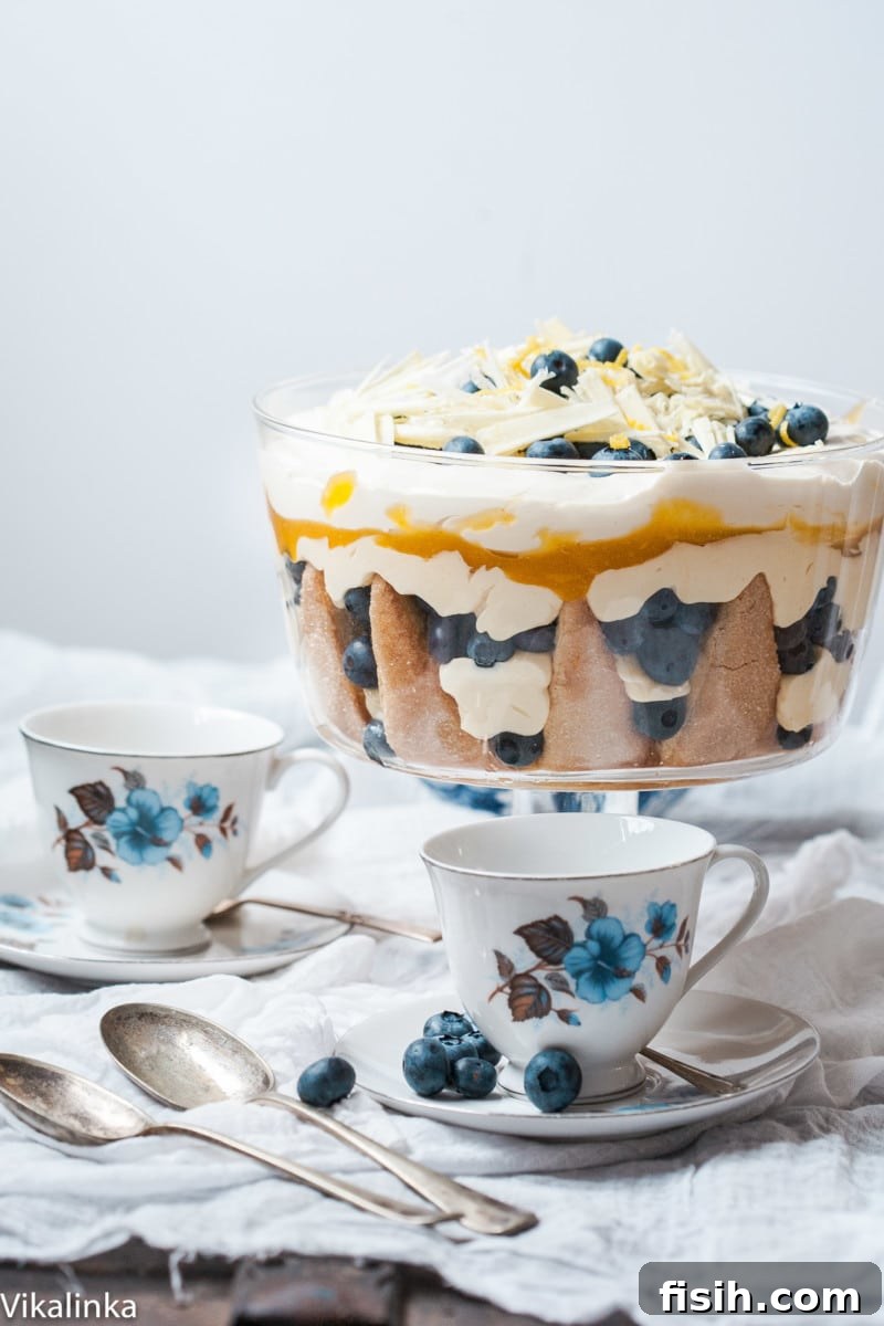 A beautifully layered lemon blueberry trifle, garnished with fresh blueberries, on a table alongside two white and blue teacups. Perfect for a summer gathering.