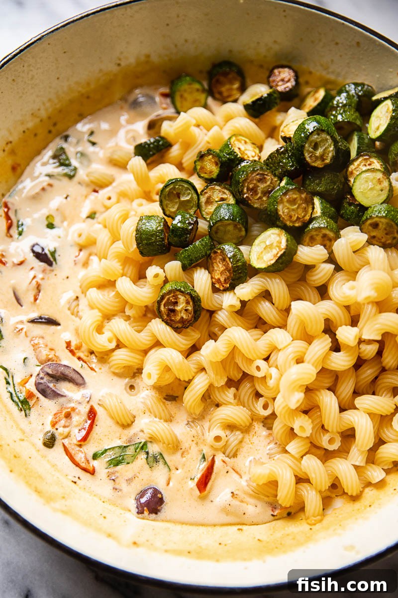 Close-up of pasta ingredients in a pan before being mixed, showing zucchini, olives, and tomatoes