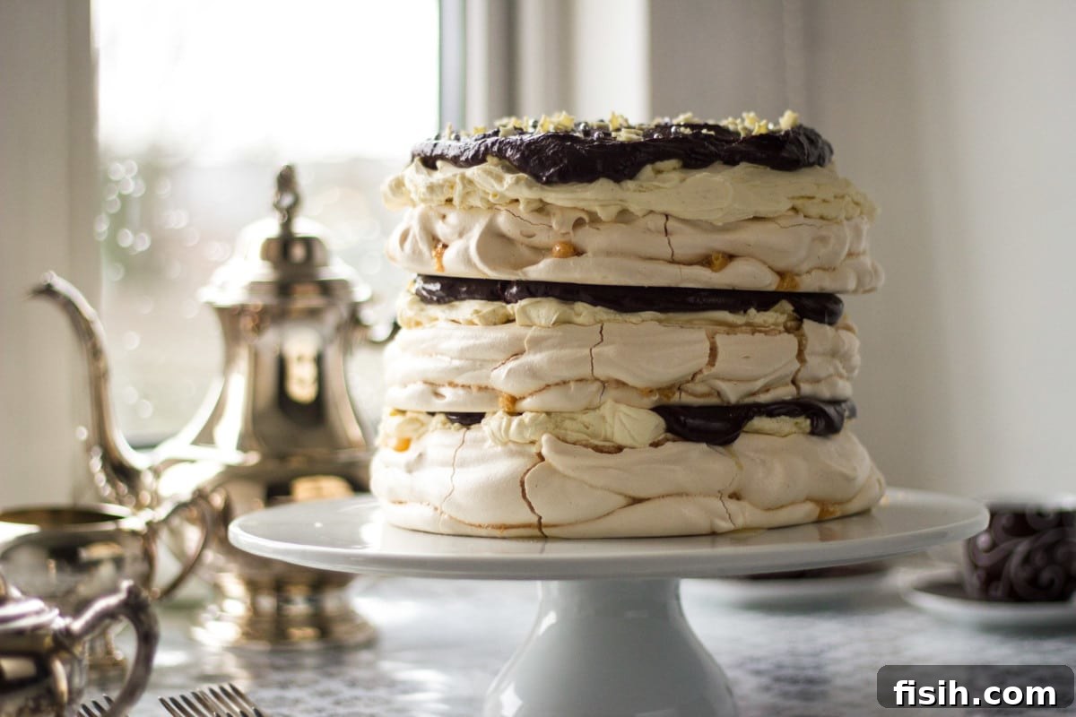 Side shot of a beautifully layered meringue cake on a white stand, with a teapot softly blurred in the background, suggesting a cozy tea-time.