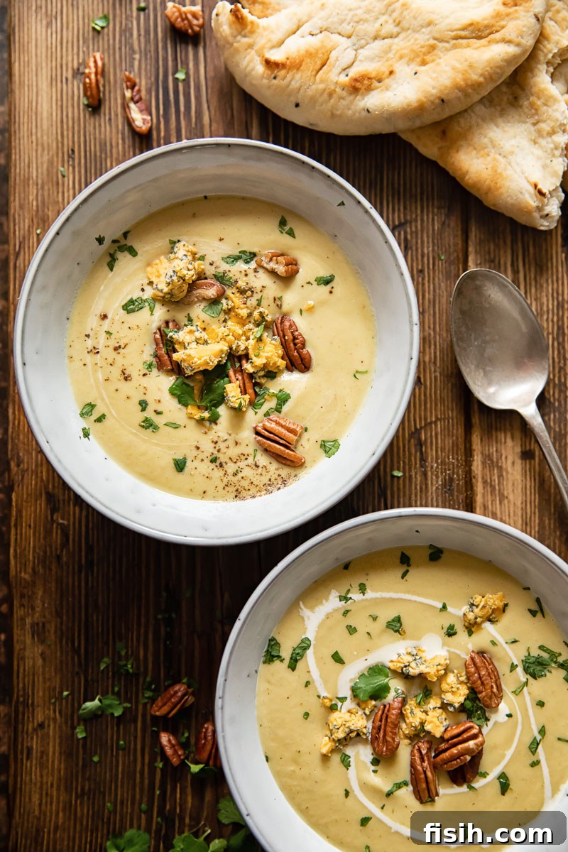 An inviting top-down view of two bowls of curried cauliflower cheese soup, garnished with crumbled Stilton and pecans, set on a warm wooden background.