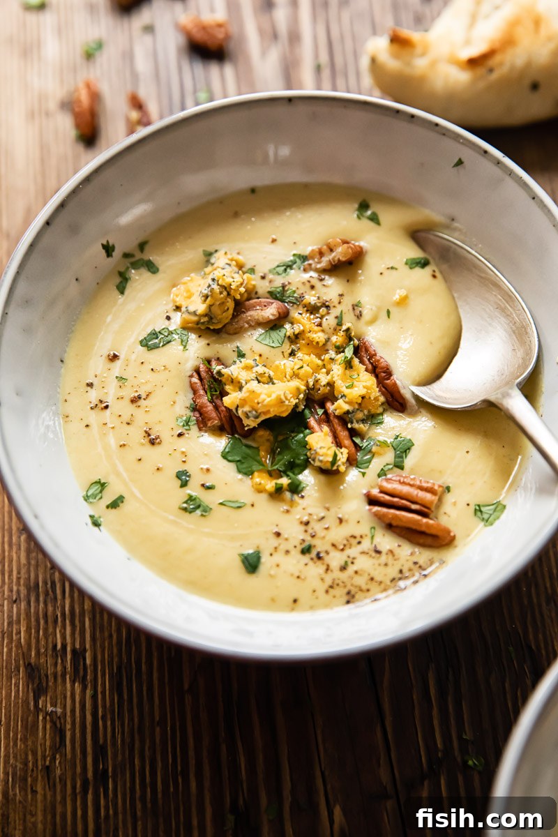 Close-up of cauliflower soup in a bowl, generously topped with crumbled Stilton, toasted pecans, and fresh herbs, highlighting its rich texture and vibrant garnishes.