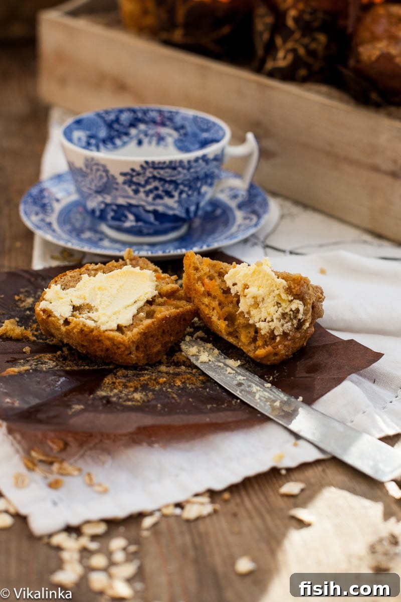 A carrot oatmeal muffin cut in half to reveal its moist, crumbly interior, served with a pat of butter and a blue teacup in the background.