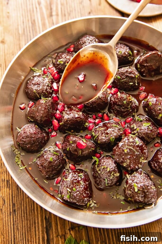 A pan filled with perfectly glazed sweet and sour meatballs, with a spoon drizzling more of the luscious pomegranate sauce over them.
