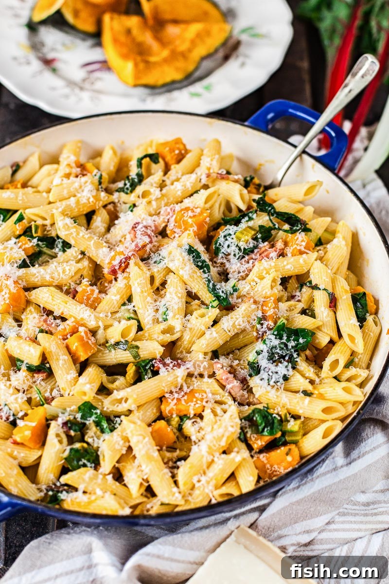 Penne pasta mixed with roasted squash and crispy bacon in a frying pan, with more roasted squash visible in the background.