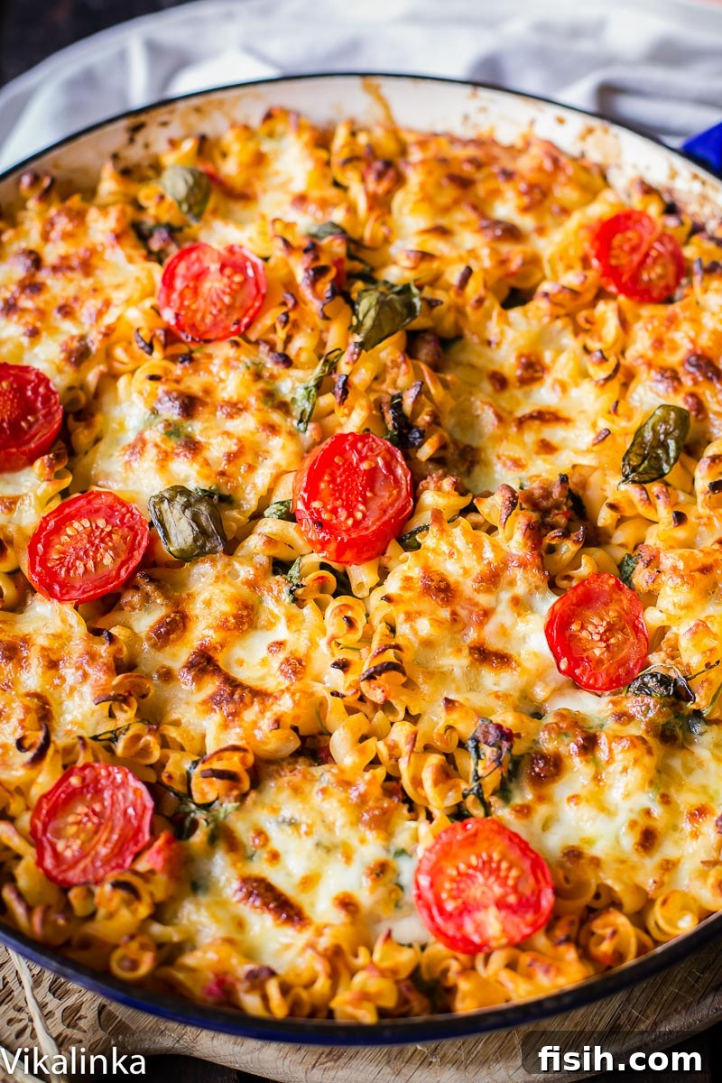 A golden-brown cheesy sausage pasta bake topped with cherry tomatoes, fresh basil, and melted mozzarella in a round baking dish, ready to serve.