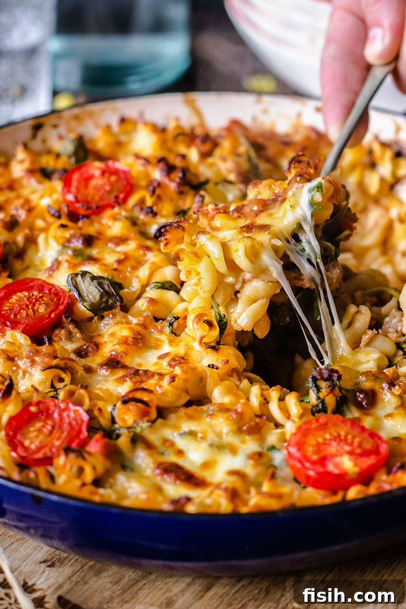 A close-up side view of a serving of the cheesy sausage pasta casserole being scooped from the pan with a spoon, showing melted cheese and rich sauce.