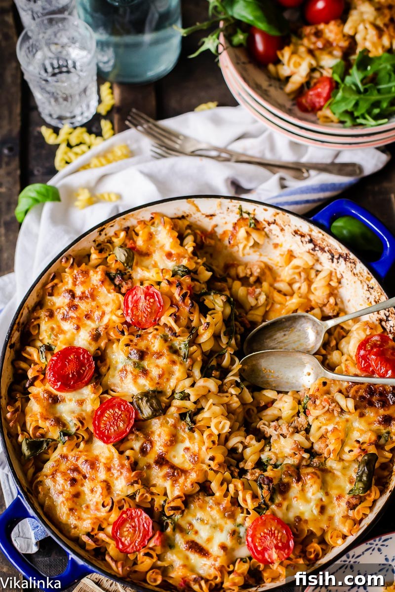 An overhead view of the completed baked pasta, generously topped with golden melted cheese, halved cherry tomatoes, and fresh basil leaves, with serving bowls and drinks blurred in the background, suggesting a ready meal.