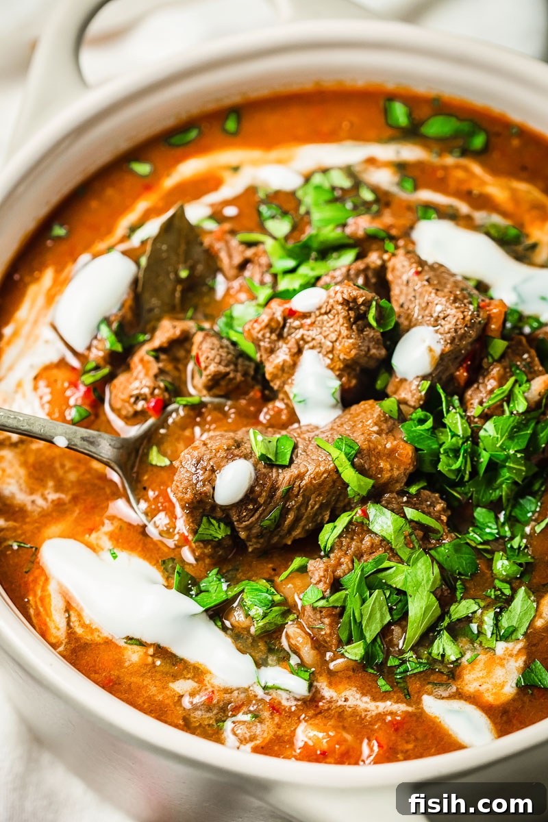 A vibrant and comforting bowl of Beef Goulash, garnished with fresh sour cream and parsley, served in a cream-colored pot.