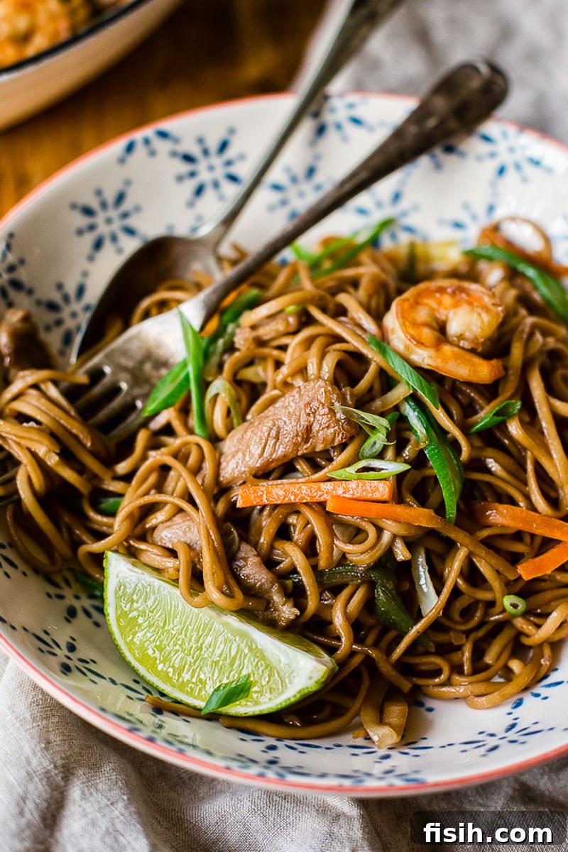 A bowl of Filipino Pancit Canton noodles with a fork, spoon, and a lime wedge on the side