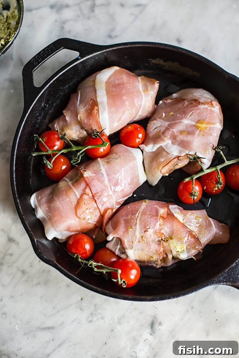 Uncooked prosciutto wrapped pesto chicken parcels on a cast iron pan with cherry tomatoes
