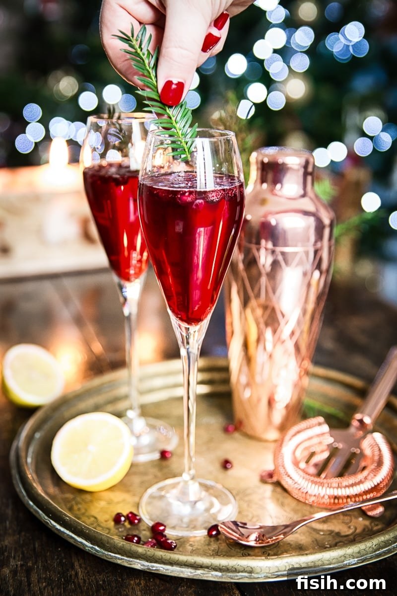 A hand gently placing a clipped pine branch garnish into a Pomegranate French 75 cocktail, alongside another ready drink