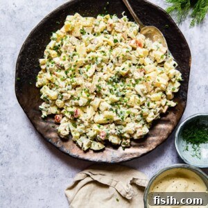 Russian potato salad on a platter with a spoon, accompanied by a cream napkin, a bowl with salad dressing, and a bowl with fresh herbs and dill.