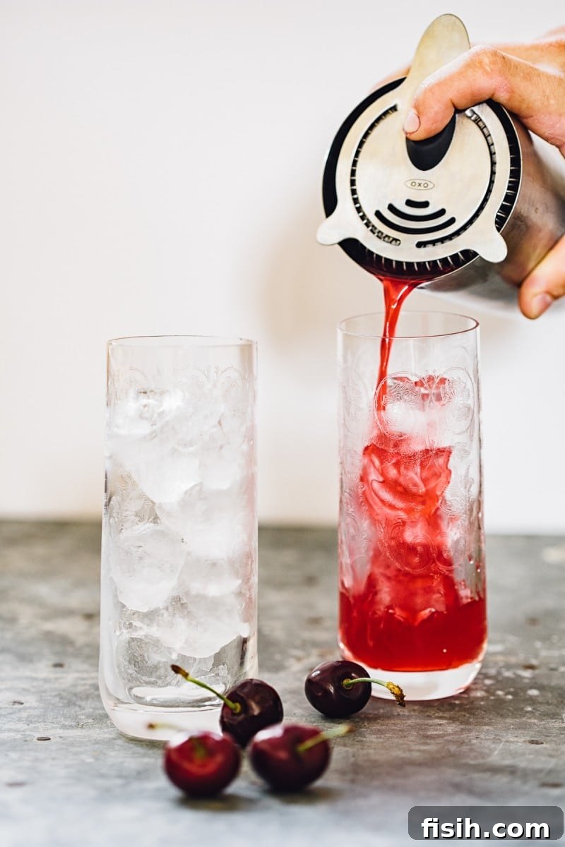 A cocktail shaker being used to pour a vibrant red drink over ice into a tall glass, with a second glass ready.