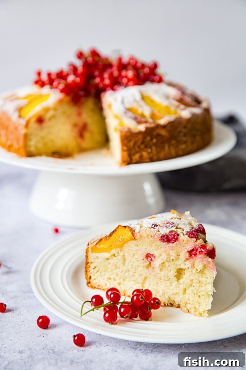 A slice of peach cake with red berries on a plate, with the rest of the cake in the background, highlighting its moist texture