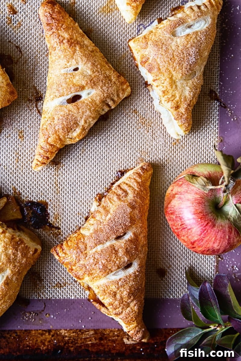Golden Apple Pockets 2 Golden-brown apple turnovers arranged beside a whole fresh apple, highlighting their rustic charm and delicious appeal.