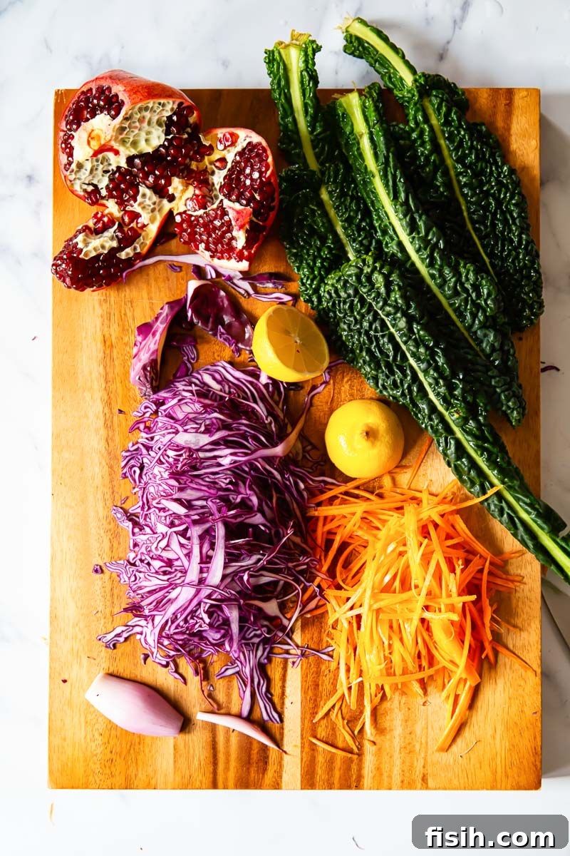 Close-up view of fresh ingredients for winter salad on a wooden cutting board: finely sliced red cabbage, vibrant green kale leaves, grated carrots, shiny red pomegranate seeds, and thinly sliced shallots, ready for preparation.