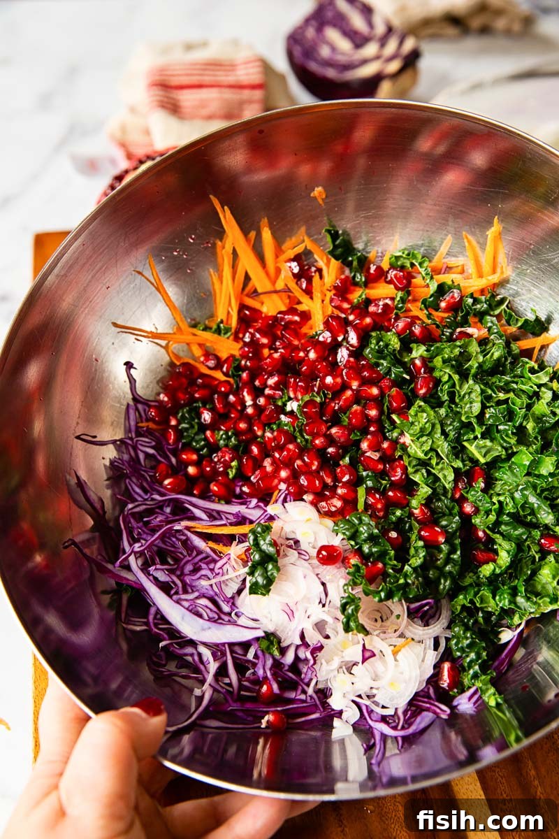 A large mixing bowl filled with freshly prepared winter salad ingredients: finely shredded red cabbage, vibrant grated carrots, tender sliced kale, and glistening pomegranate seeds, ready for the dressing.