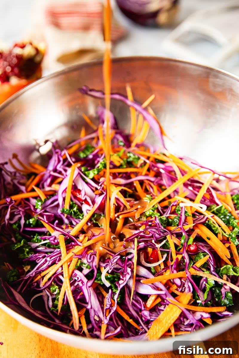 Overhead shot of a vibrant red cabbage, kale, and pomegranate salad in a bowl, with a zesty vinaigrette being drizzled generously over the fresh ingredients, ready to be tossed.