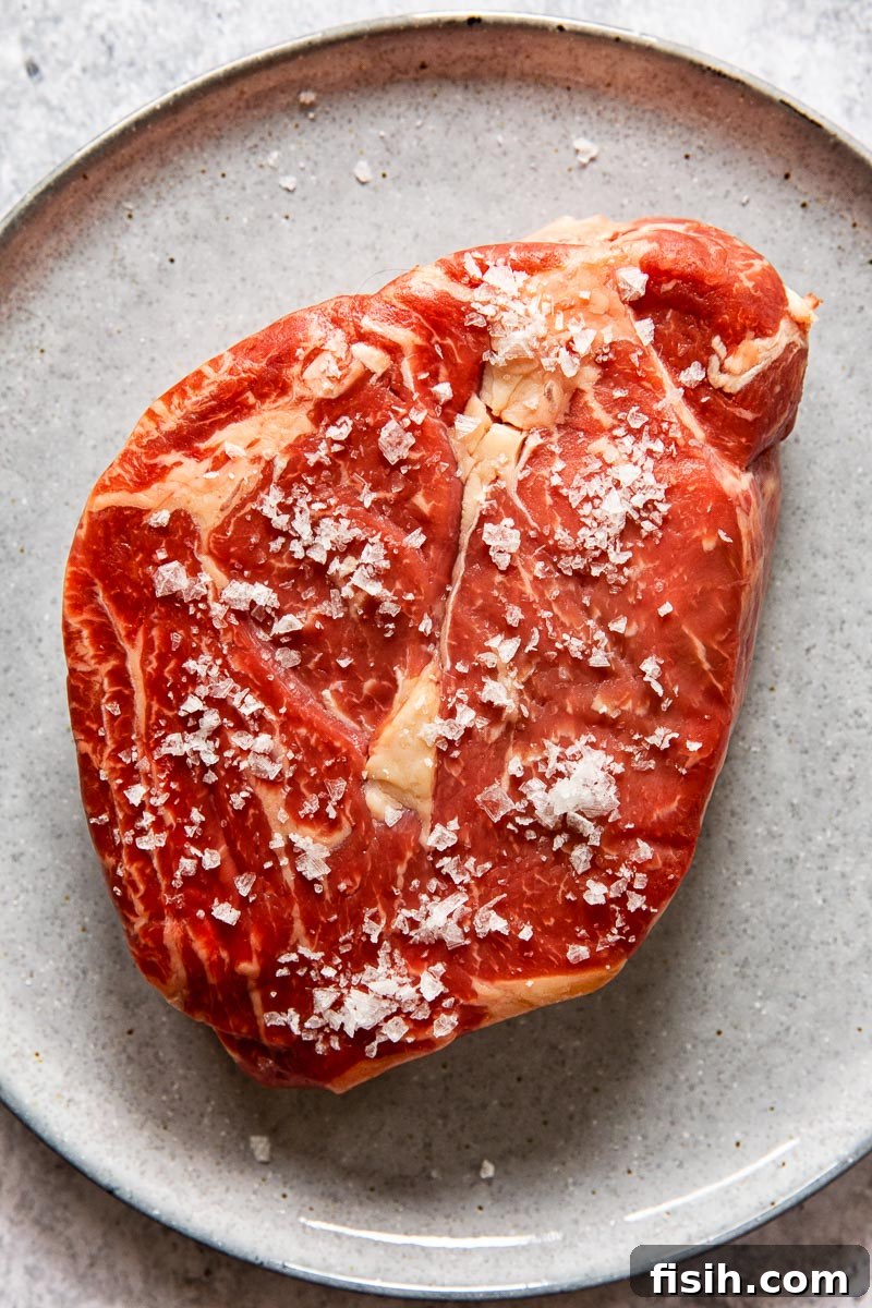 Raw ribeye steak, generously sprinkled with coarse flaked salt, resting on a dark grey plate.