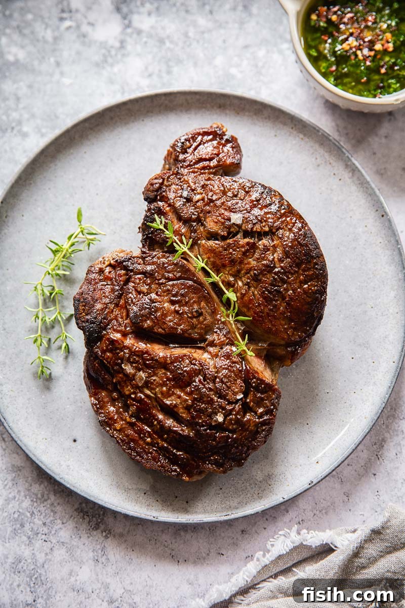 Perfectly cooked ribeye steak resting on a grey plate, with a small bowl of green herb sauce nearby.