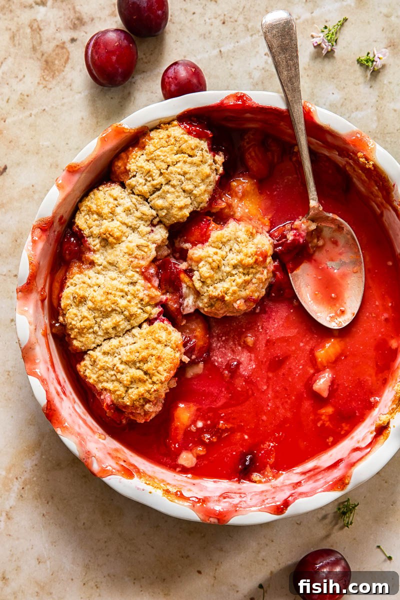 A round baking dish with a partially eaten plum cobbler, showing the golden topping and juicy fruit underneath, with a spoon resting beside it.