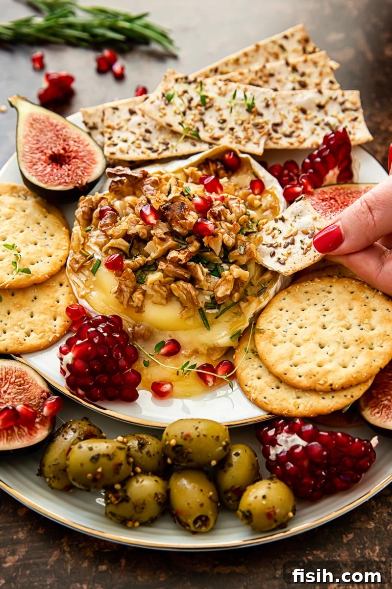 Baked brie topped with walnuts and pomegranate seeds, crackers, figs and olives around the plate.