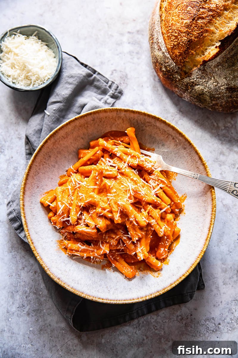 Velvety Vodka Pasta Sauce 5 A top-down view of a bowl of pasta alla vodka, accompanied by a fork, a slice of crusty bread, and grated Parmesan cheese in a small blue bowl.