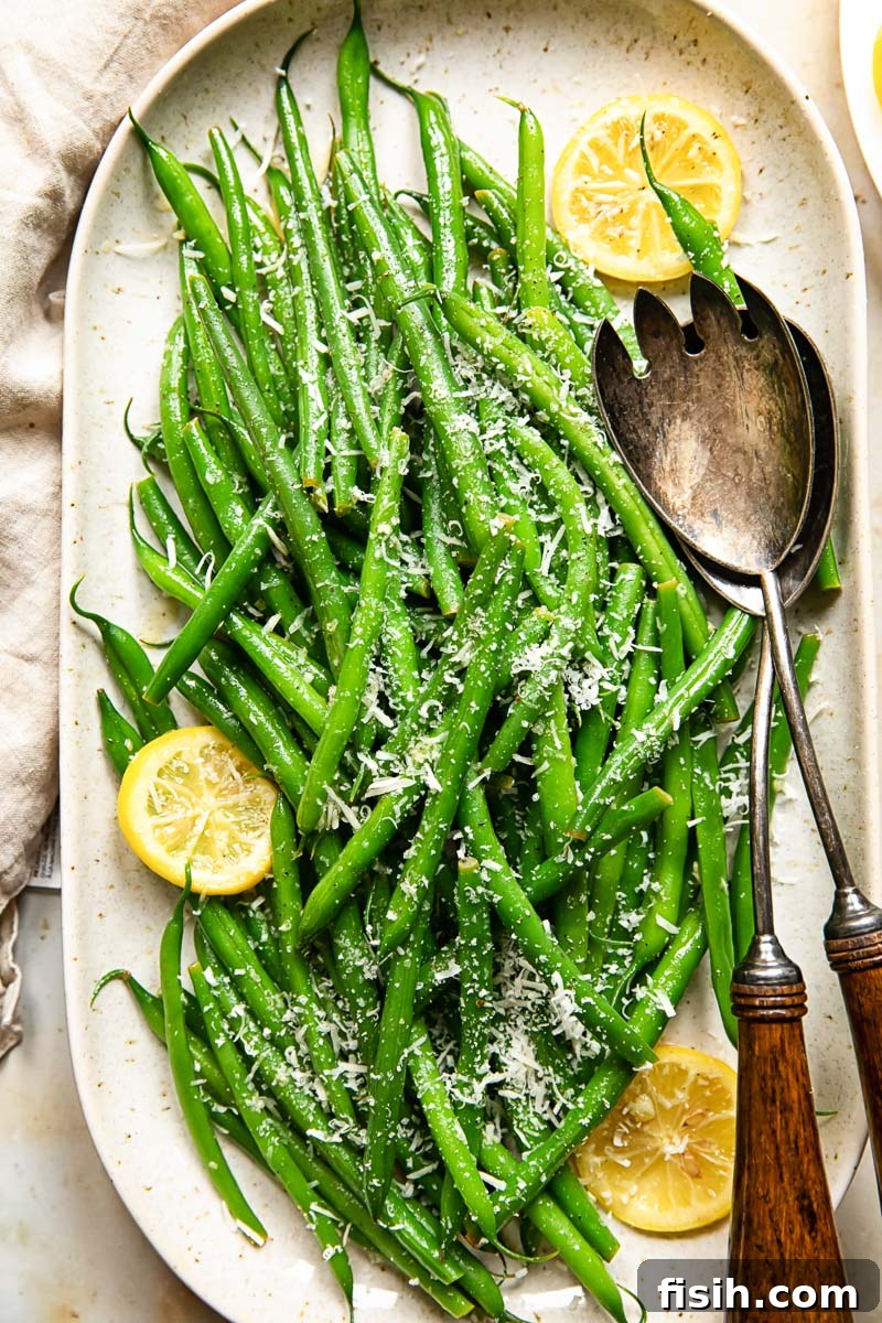 Bright Lemon-Garlic Parmesan Green Beans 5 Top-down view of vibrant green beans garnished with fresh lemon slices and a generous dusting of Parmesan cheese on a white platter.