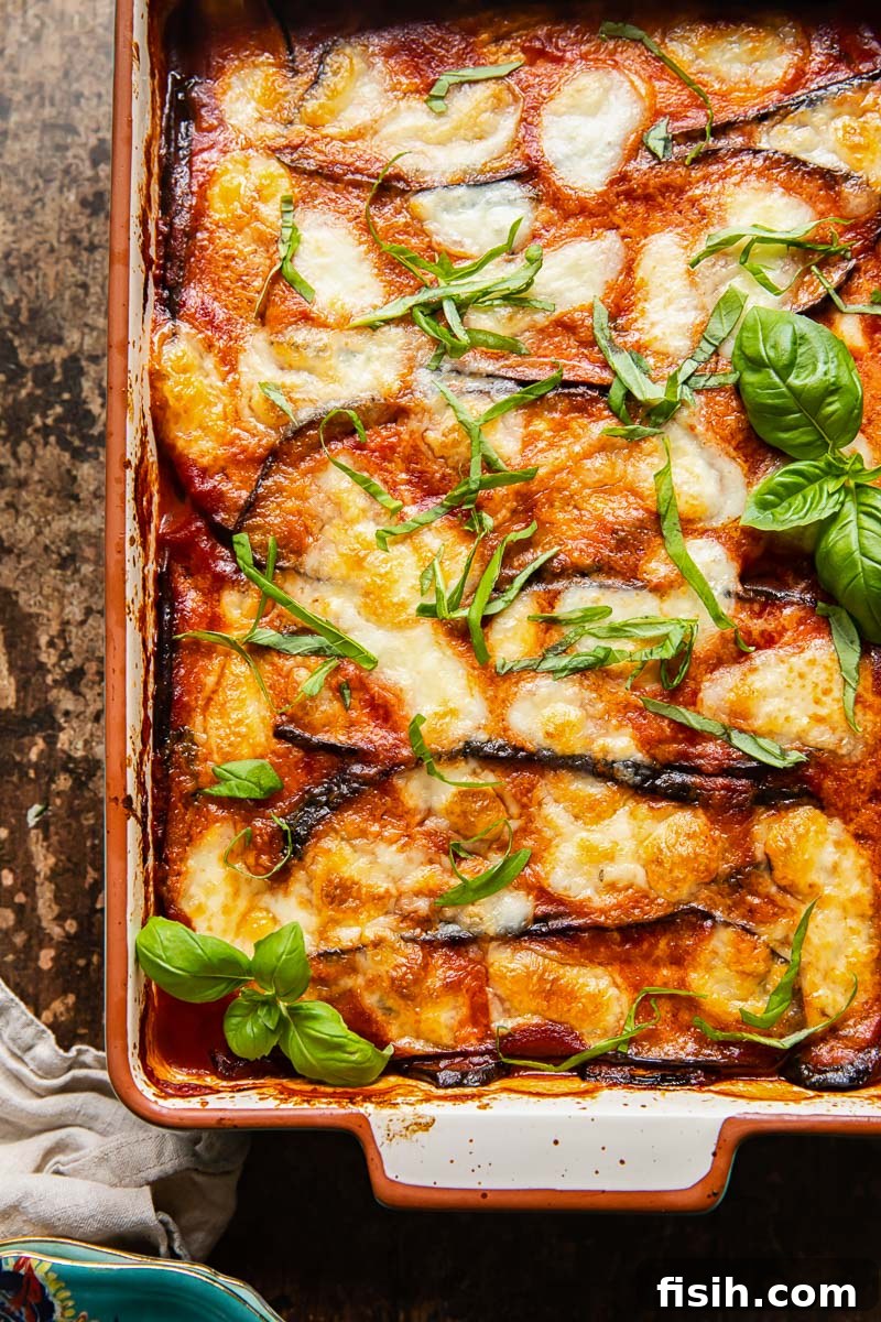 Guilt-Free Eggplant Parmigiana 2 Top-down view of glistening, layered Eggplant Parmesan in a rectangular baking dish, artfully placed on a rustic dark wooden background, garnished with fresh basil leaves.