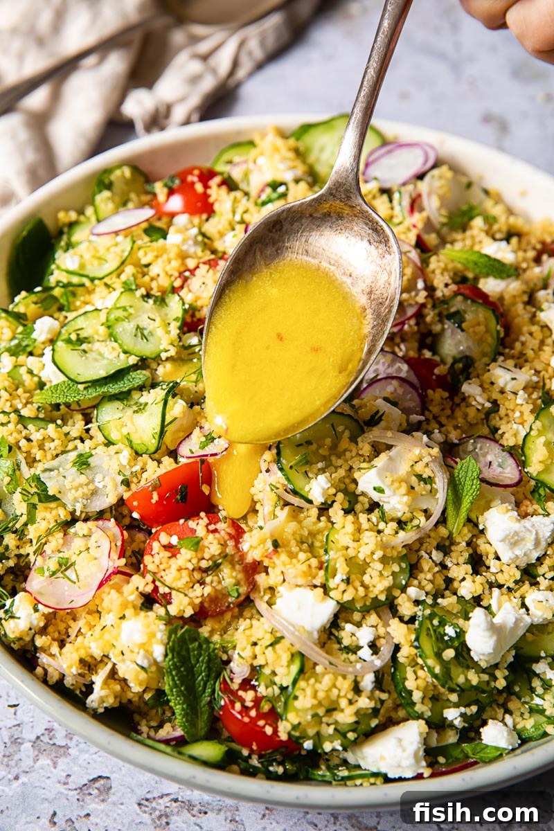 A spoon with a vibrant yellow lemon salad dressing being elegantly drizzled over a fresh couscous salad, highlighting the texture and colors of the dish.