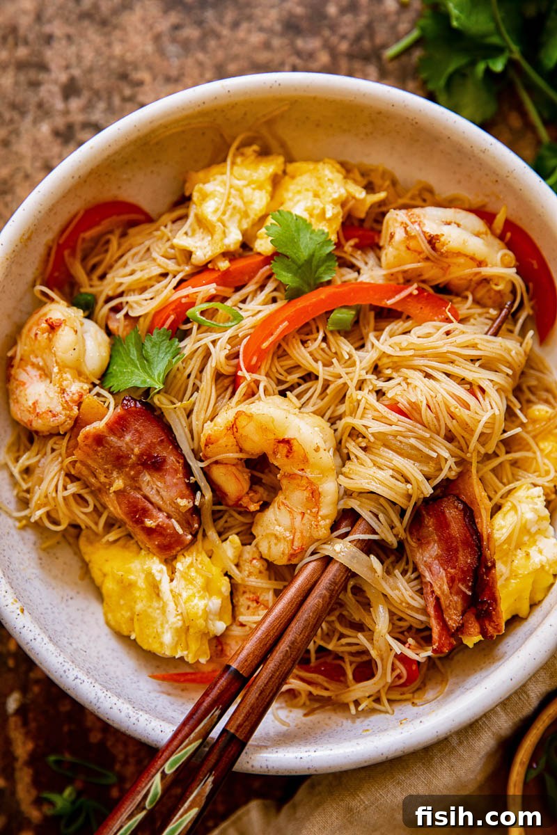 Close-up of stir-fried Singapore Noodles with prawns and red peppers in a ceramic bowl, with chopsticks resting on the side