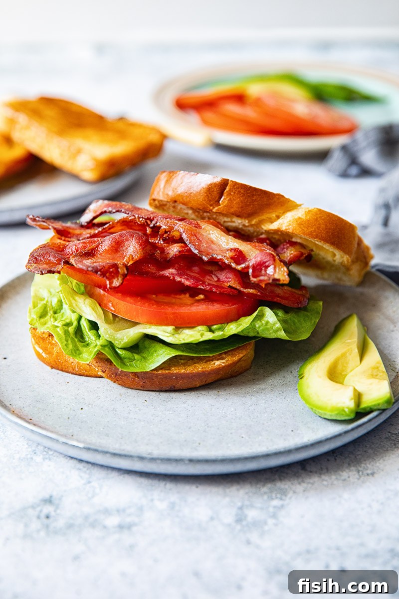 Side view of a perfectly assembled bacon, lettuce, and tomato sandwich on a grey plate, with a half-sliced avocado positioned next to it.