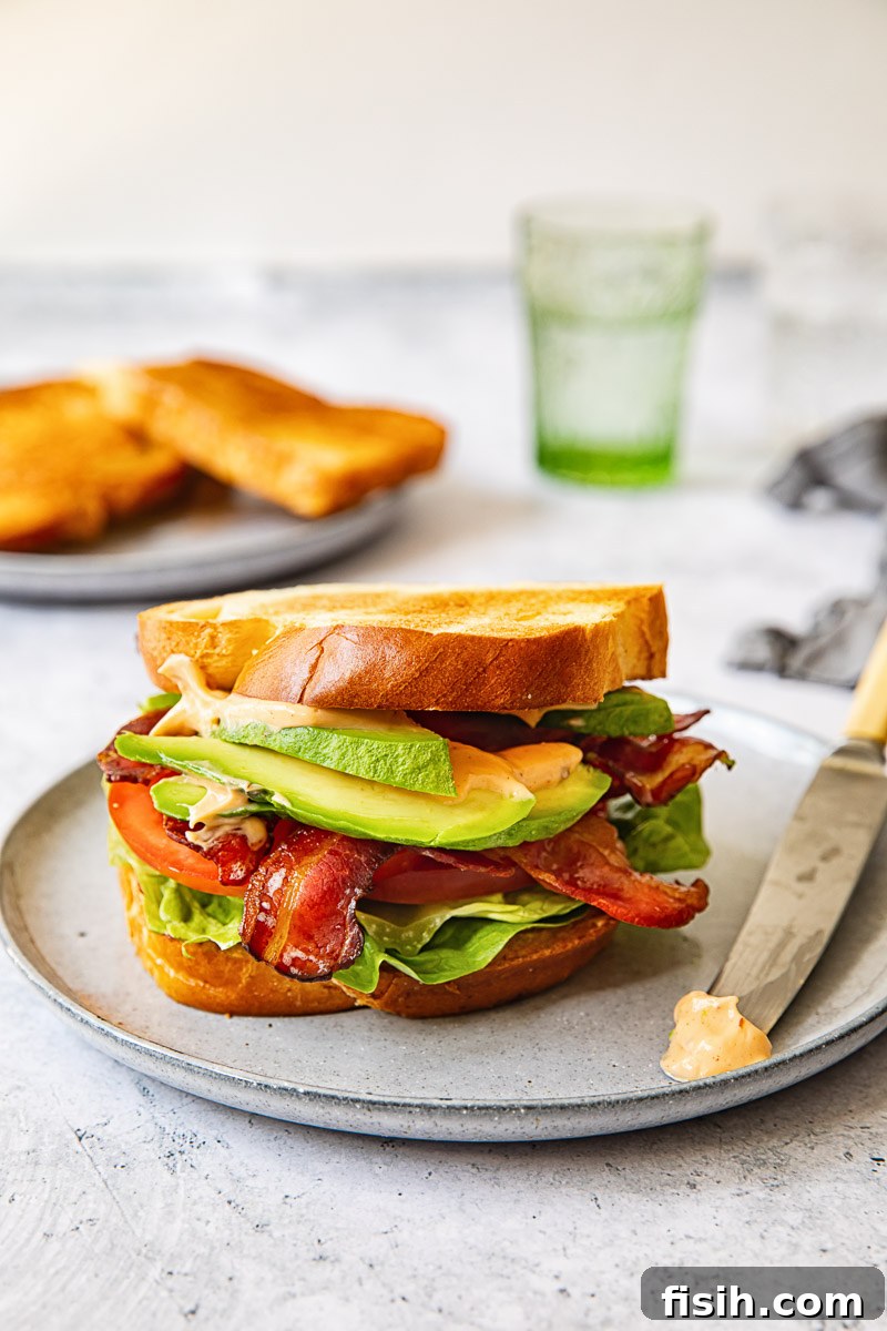A complete Maple BLT sandwich resting on a grey plate, with a butter knife beside it and a glass of green water in the soft-focus background.
