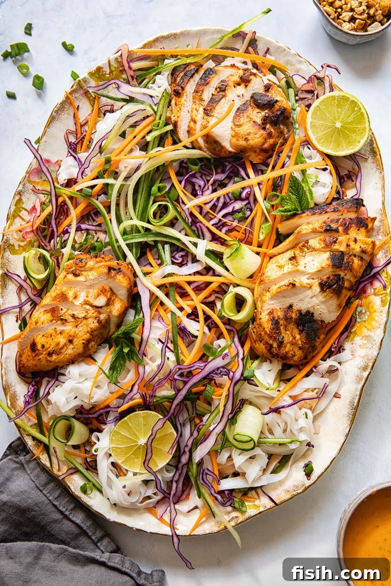 A large oval platter showcasing a vibrant Thai grilled chicken salad, featuring sliced chicken breasts, colorful vegetables, and rice noodles drizzled with peanut sauce.