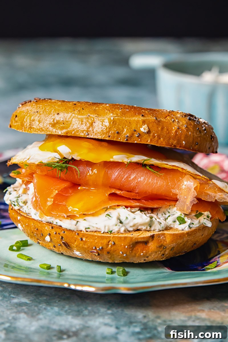 A beautifully assembled breakfast bagel sandwich with herbed cream cheese, smoked salmon, and a fried egg with a runny yolk, served on a whole grain bagel.