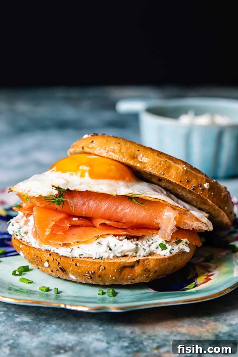 A close-up of the breakfast bagel sandwich, showing the layers of herbed cream cheese, smoked salmon, and a perfectly cooked fried egg on a toasted bagel.