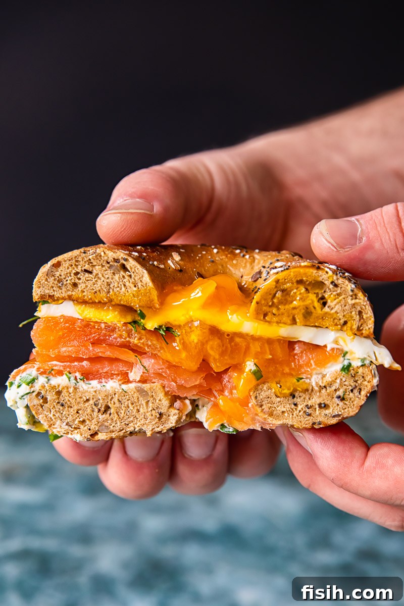A stunning cross-section view of the breakfast bagel sandwich, showcasing the vibrant layers of toasted bagel, herbed cream cheese, folded smoked salmon, and a perfectly cooked fried egg with a glistening runny yolk.