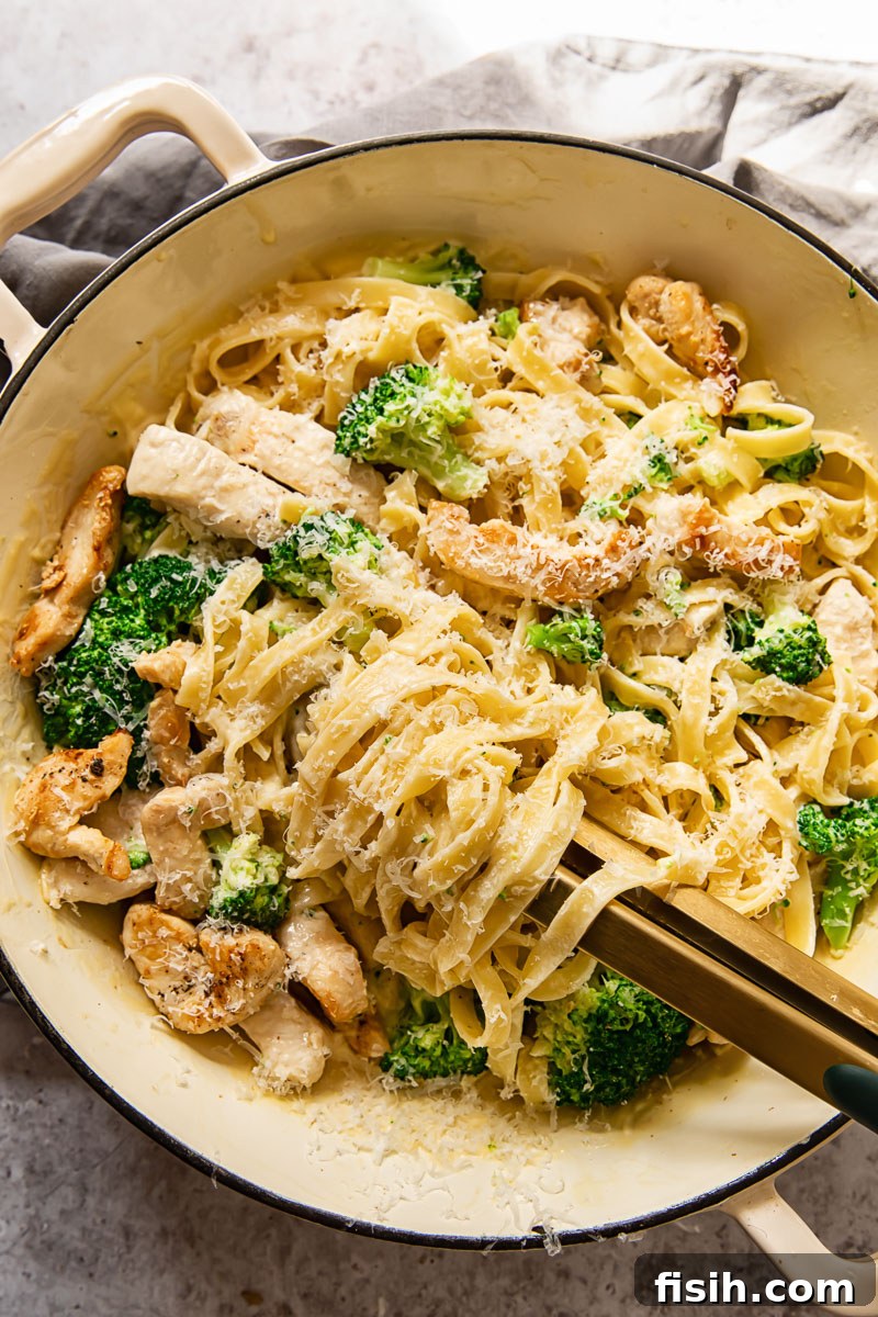 top down view of chicken broccoli alfredo fettuccine in a pan with tongs in it, ready to be served