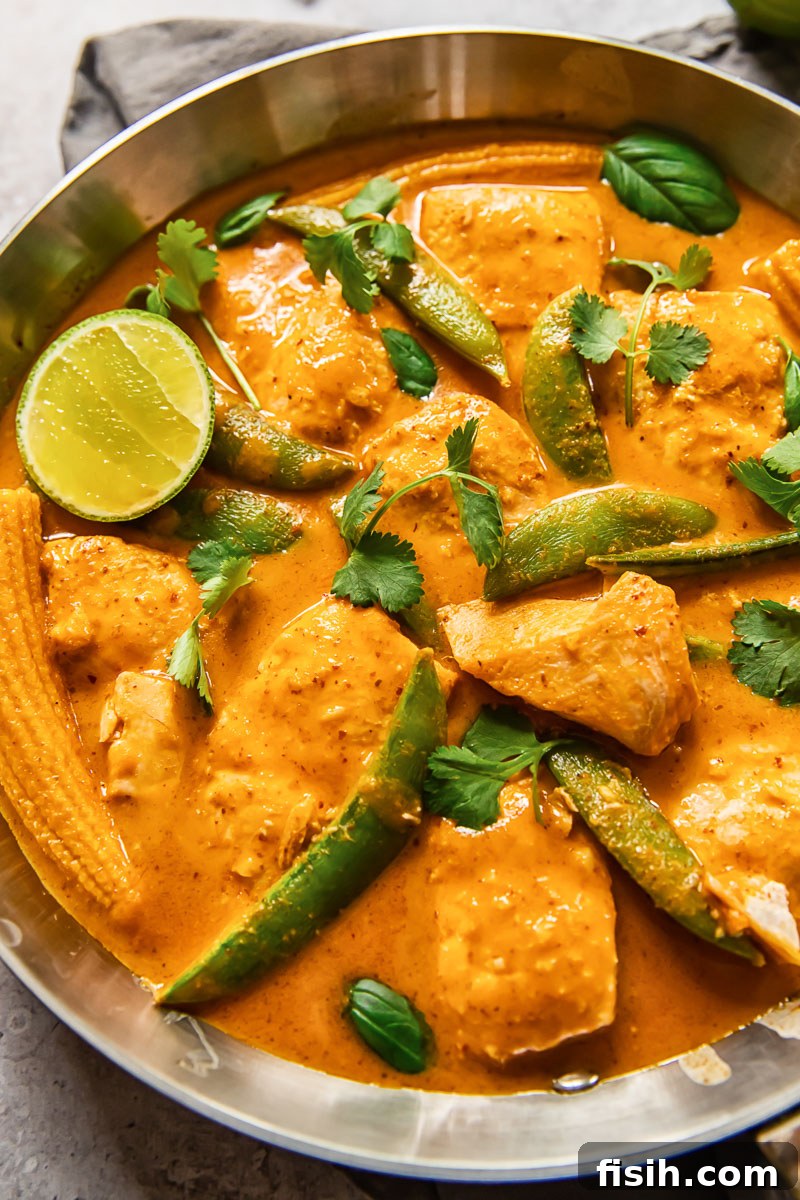A vibrant red salmon curry cooking in a pan, filled with tender fish, sugar snap peas, and baby corn.