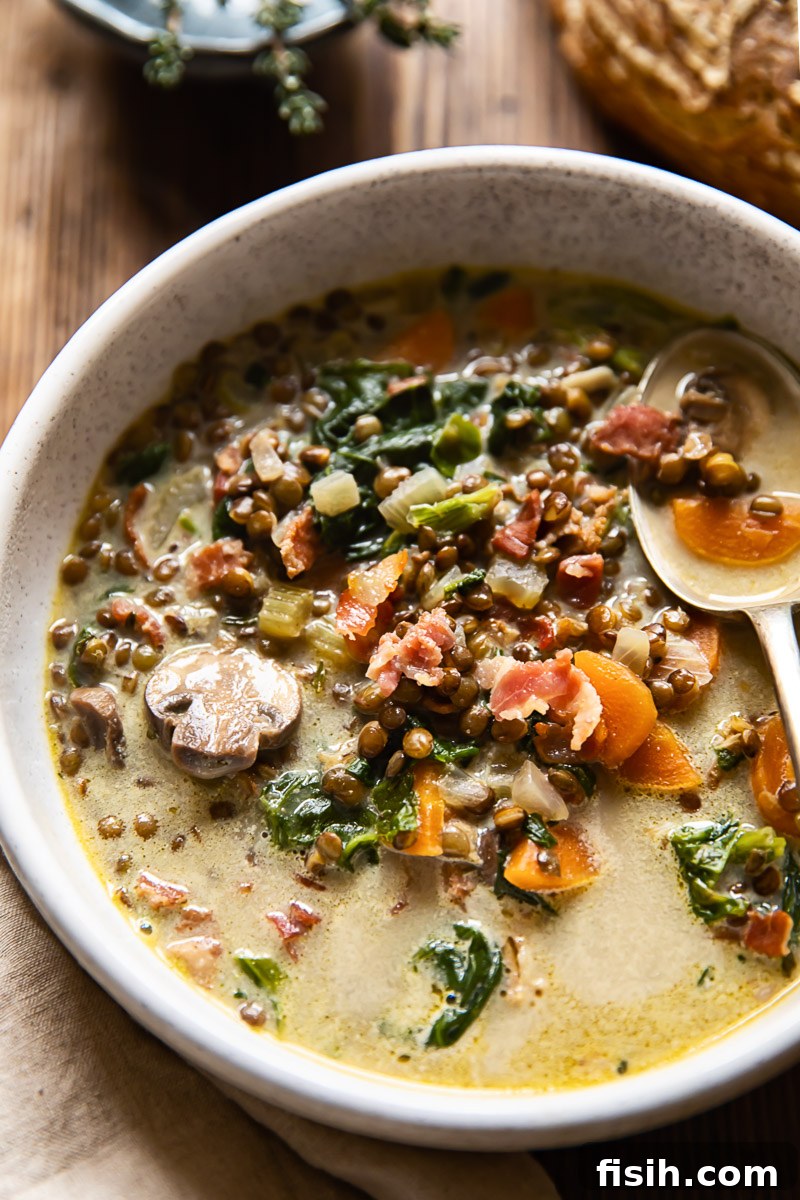 A beige bowl brimming with steaming vegetable, bacon, and lentil soup, ready to be enjoyed.