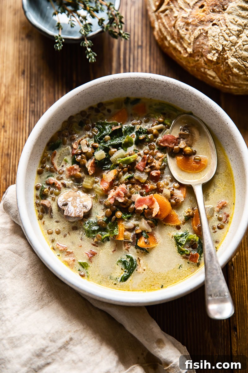 An inviting top-down view of a bowl of lentil and bacon soup nestled beside a rustic loaf of bread on a wooden table, emphasizing its cozy appeal.