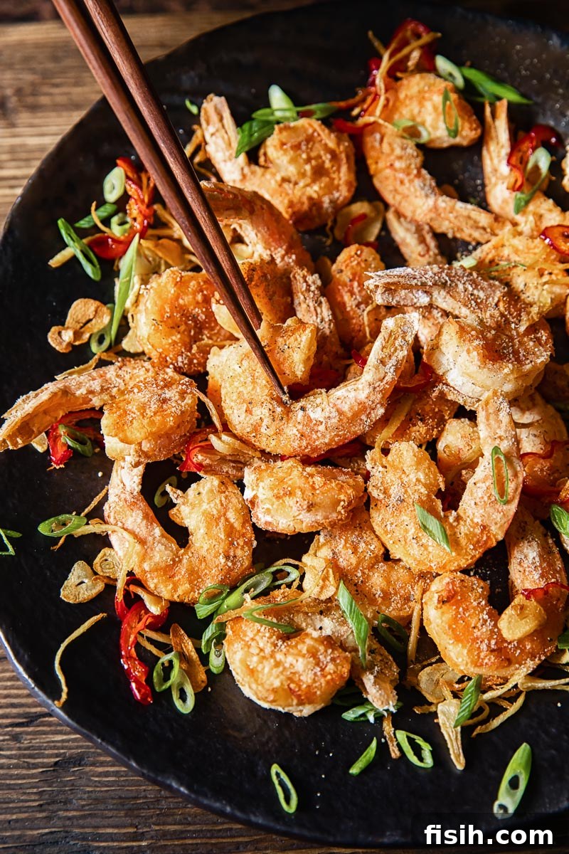 Crispy Air Fryer Salt and Pepper Shrimp 6 Top-down view of a dark platter filled with golden Salt and Pepper Prawns, with one shrimp being delicately picked up by chopsticks