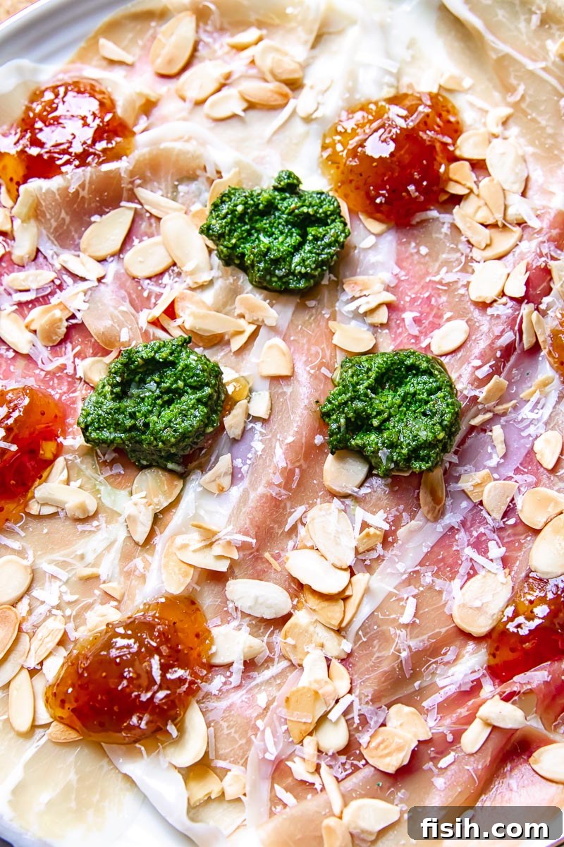 Close-up detail of an Italian charcuterie board, highlighting the textures of prosciutto, the bright green pesto, and the rich fig jam.