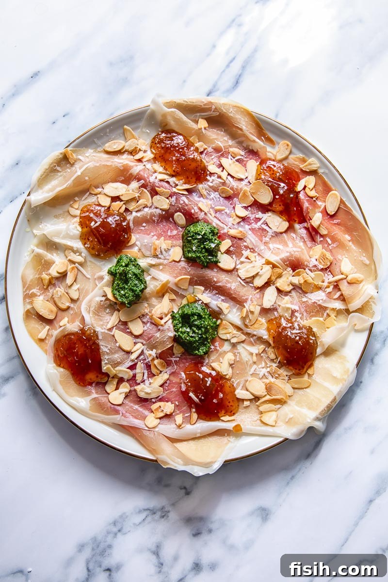 Overhead shot of a vibrant prosciutto platter, artfully adorned with dollops of bright green pesto, rich fig jam, and crunchy toasted almonds.