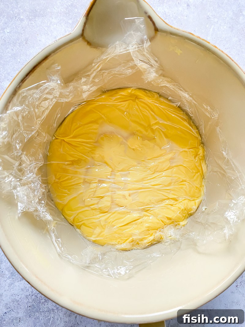Top-down view of creamy custard in a bowl covered with plastic wrap to prevent skin formation