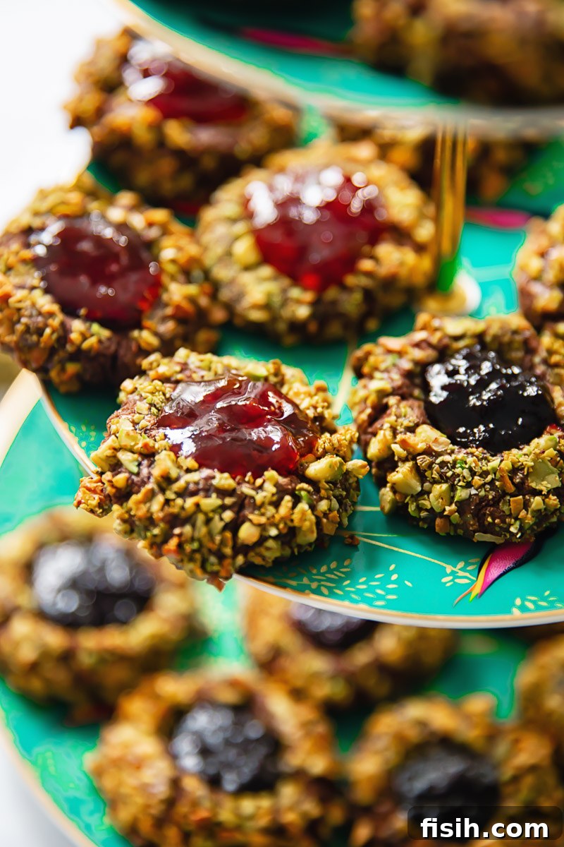 Close-up view of a pile of chocolate thumbprint cookies, generously coated in chopped pistachios and filled with glistening jam, ready to be enjoyed.