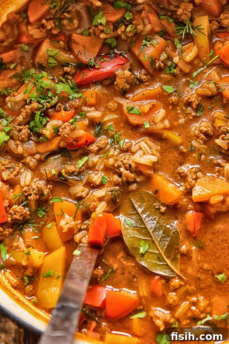 Close-up shot of hearty unstuffed pepper soup simmering in a large pot, with ground beef and rice visible, and a ladle resting inside.
